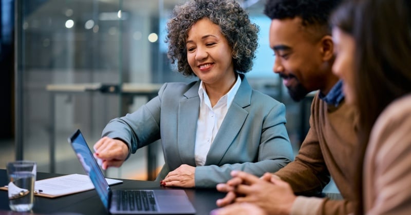 Professional advisor smiling while presenting financial data on a laptop to two clients during a business meeting in a modern office setting.