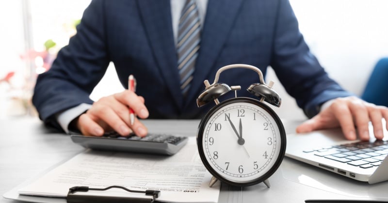 Business professional in a suit working at a desk with a calculator and laptop, with a large analog alarm clock in the foreground showing the time as 10:10.