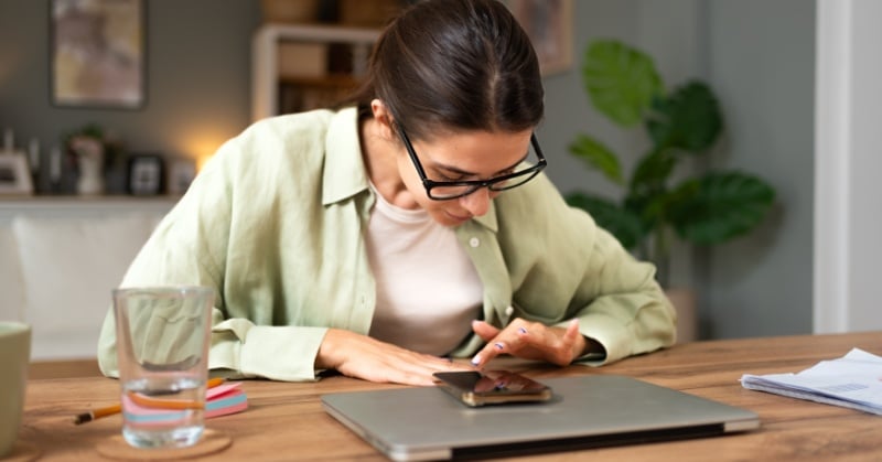A woman wearing glasses and a light green shirt concentrates on her smartphone while seated at a desk with a laptop, glass of water, and scattered papers.