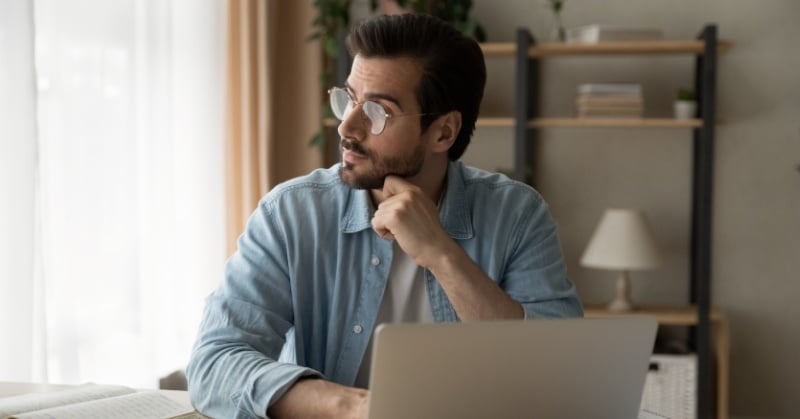 A bearded man in glasses and a denim shirt sits at a desk with a laptop, gazing thoughtfully out the window with his chin resting on his hand.