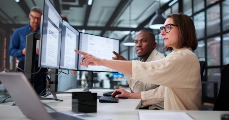 Professionals reviewing documents on multiple monitors in an office setting.