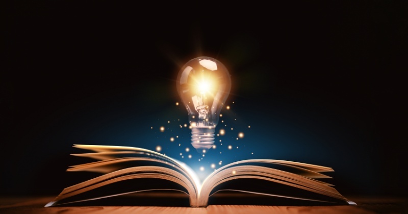 An open book on a wooden surface with its pages fanned out, emitting glowing light particles. Above the book hovers an illuminated light bulb, symbolizing knowledge, creativity, or inspiration, set against a dark background.