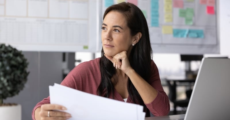 Woman seated at a desk in an office, holding papers and looking thoughtfully to the side beside a laptop.