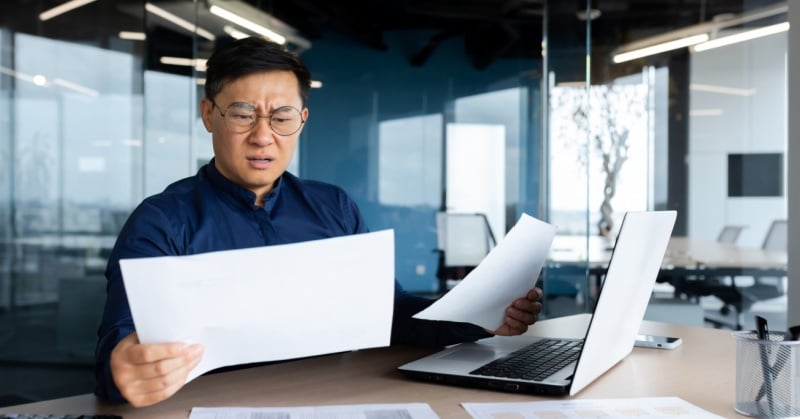 A man wearing glasses and a dark blue shirt sits at a desk in a modern office, looking perplexed as he holds and examines two sheets of paper, with an open laptop in front of him.