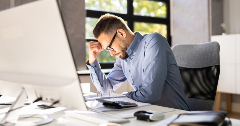 Man working at a desk with a computer and calculator, appearing stressed while reviewing accounting paperwork in an office.