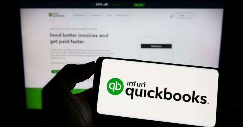 A smartphone displays the Intuit QuickBooks logo with the QuickBooks website blurred in the background, highlighting the platform's invoicing features.