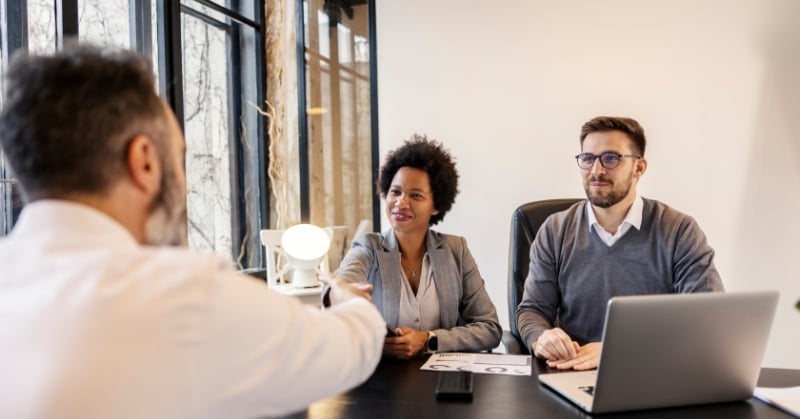 Two accounting professionals meet with a client in a modern office, smiling and shaking hands across a desk with documents and a laptop.