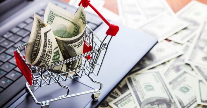 Shopping cart filled with U.S. dollar bills sitting on a laptop, with additional cash scattered in the background.