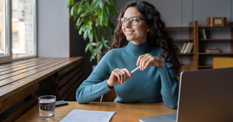 A woman with long curly hair and glasses, wearing a teal sweater, smiles while sitting at a desk with a laptop, paperwork, and a glass of water, gazing out of a window in a modern office setting.