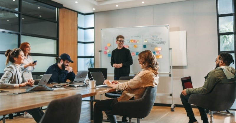 A group of professionals collaborates in a bright, modern office, with one person leading a discussion at a whiteboard covered in colorful notes.