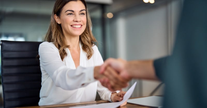 Smiling woman in professional attire shakes hands with an unseen person across a desk, holding a resume during a job interview in a modern office setting.