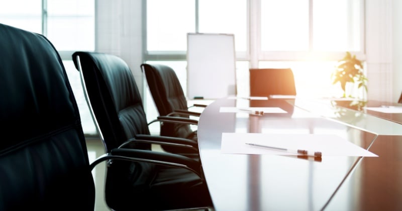 A modern, sunlit corporate boardroom with empty black leather chairs arranged around a polished conference table. Papers and pens are placed neatly at each seat, with a flipchart and potted plant visible in the background.