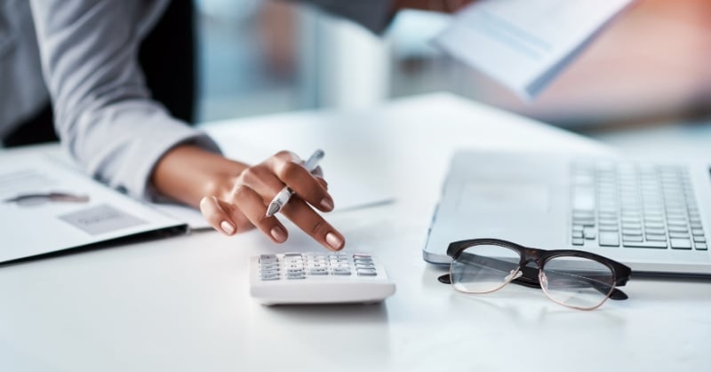 A professional accountant's hand using a calculator at a modern white desk, with documents, eyeglasses, and a laptop nearby, symbolizing financial analysis or payroll processing work.