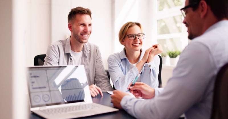 A smiling couple meets with a financial advisor in a bright office, with a laptop displaying charts on the desk.