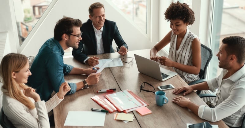 Diverse group of professionals collaborating around a conference table with laptops, documents, and coffee, representing teamwork in a modern office.