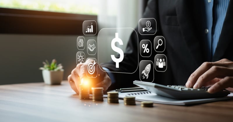 A business professional calculates finances at a desk with a digital dollar sign and finance-related icons floating above stacked coins, symbolizing financial management and strategy.