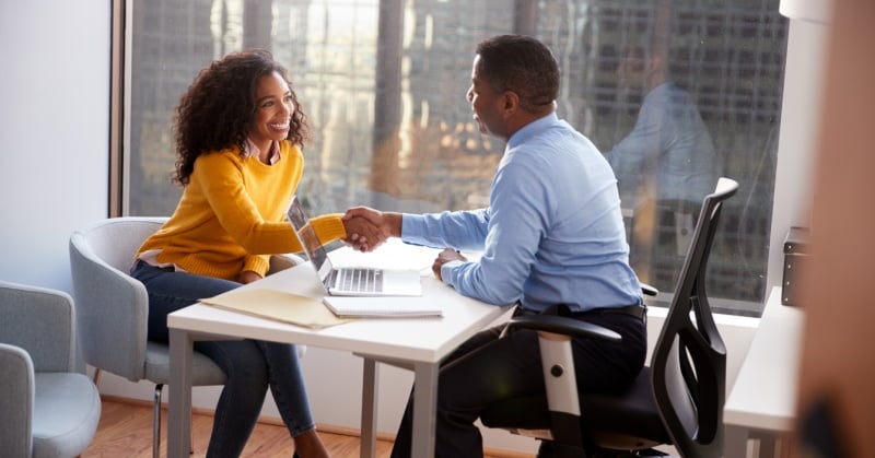 A woman and a man smile and shake hands across a desk during a professional meeting in a modern office with large windows.
