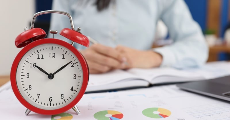 A close-up image shows a red alarm clock reading 9:50 placed on a desk covered with charts and graphs, with a person in business attire working in the background, suggesting a focus on time management and productivity.