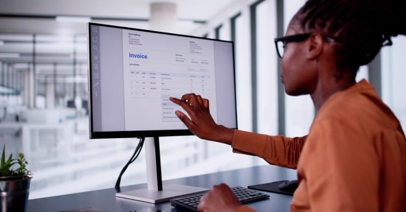 A professional woman wearing glasses and a brown blouse interacts with an invoice on a computer screen in a modern office setting.