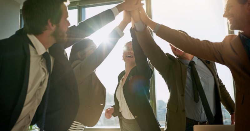 Group of five business professionals smiling and high-fiving in a brightly lit office, celebrating teamwork and success.