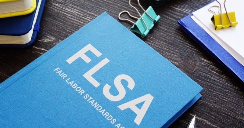 blue book that says FLSA Fair Labor Standards Act on a brown wood desk with clips and other office accessories strewn about