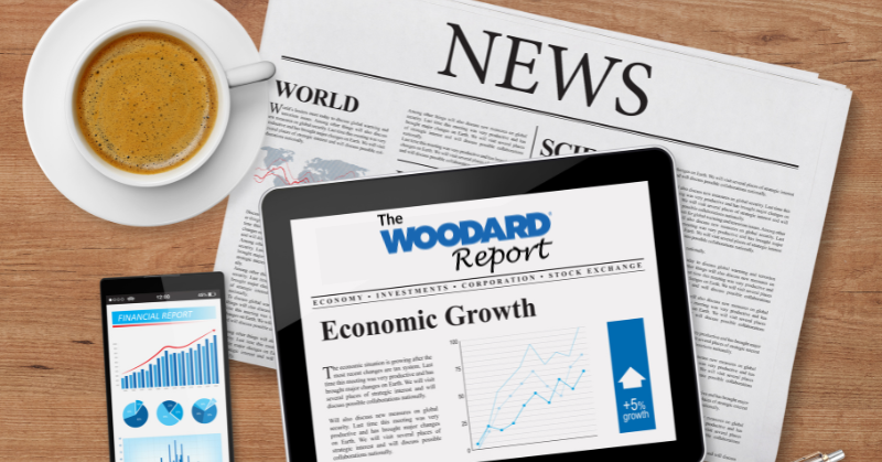 Overhead view of a tablet displaying a Woodard Report economic growth article beside a smartphone with charts, resting on a newspaper-covered wooden desk with a cup of coffee.