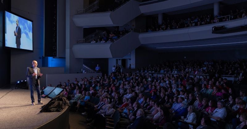 Joe Woodard delivering a keynote address at Scaling New Heights, speaking on stage to a large audience of accounting professionals in a modern auditorium.