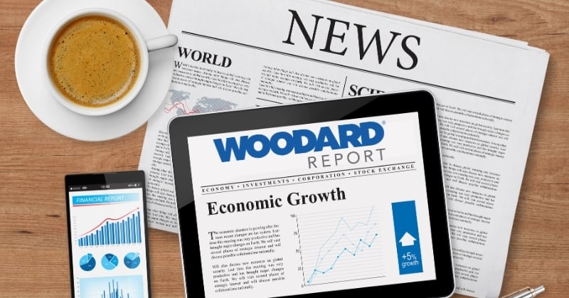 A tablet displaying The Woodard Report on economic growth sits on top of a newspaper on a wooden desk, next to a cup of coffee and a smartphone showing financial charts.