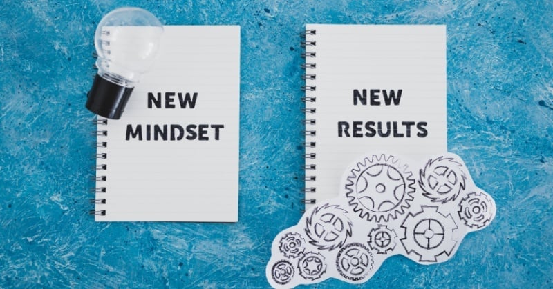 Two notebooks on a blue textured surface—one labeled "NEW MINDSET" with a lightbulb beside it, and the other labeled "NEW RESULTS" with illustrated paper gears beneath it—symbolizing the connection between mindset and outcomes.