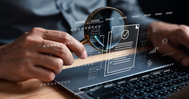 laptop with business tech holograms floating above it on desk human seated next to it holding magnifying glass up to keyboard