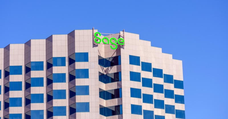 A modern office building with a tan and gray facade and blue-tinted windows. At the top of the building is a bright green “Sage” logo, representing the accounting and business software company, against a clear blue sky.