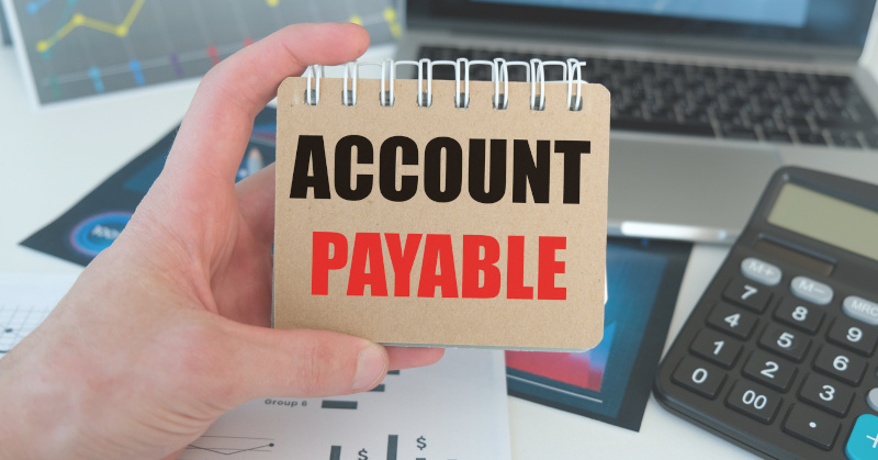 accounts payable written on beige notepad held up by human hand above a desk with reports on it