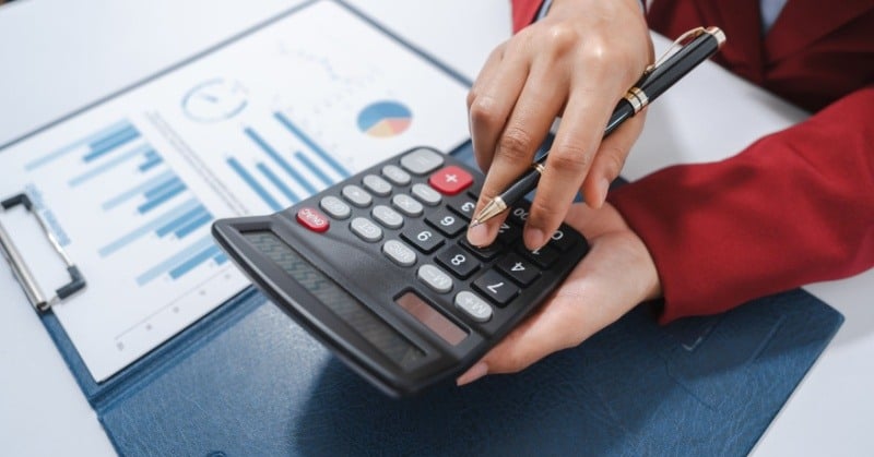 person in a red blazer holding a pen and a calculator displaying the number 9999, with financial charts visible on a clipboard on a desk