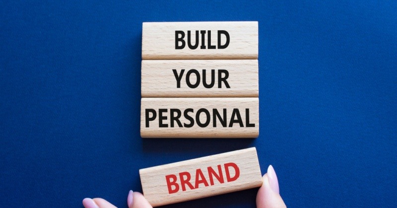 blue background light brown wood rectangular blocks stacked on top of each other with black text and one with red text saying BUILD YOUR PERSONAL BRAND being put in place by human hand with pink manicured nails