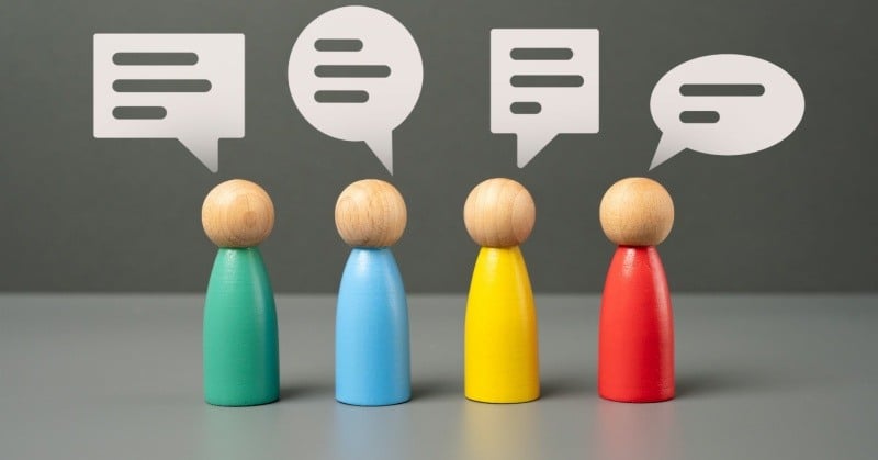 Multi-colored pegs representing multiple people on a gray table with gray background with white speech bubbles above their heads in different shapes