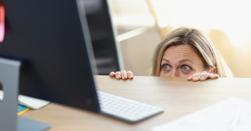 Woman kneeling under light brown desk peering over edge eyes and fingers visible looking at computer screen sitting behind keyboard