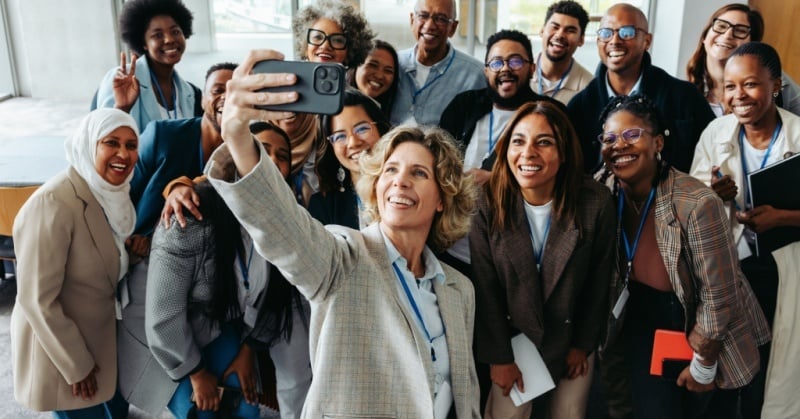 Group of diverse professionals smiling and posing for a selfie at a conference, wearing name badges and business attire in a bright, modern venue.