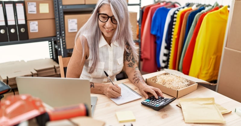middle age gray-haired woman glasses white blouse surrounded by boxes shelves and clothes in various colors working with calculator and laptop business ecommerce