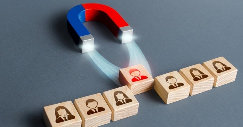 Red and blue magnet pulls a red person figure out of the row of wooden blocks with similar figures, recruiting participants from other company