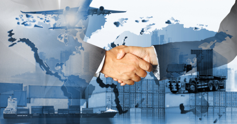 Closeup of two people's hands shaking wearing dark business suits in front of a graphjc depicting a truck, a cargo ship and shipping containers, a dock, a plane, a city's skyline, and a graphic of each continent on the globe faded together, largely colored light to dark blue except for the hands which are colored realistically