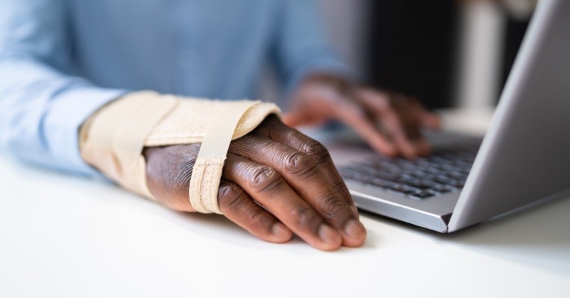 Closeup of the hands of someone seated at a white tabletop with a blue shirt typing on a laptop with a brace around their right hand in the foreground background slightly blurry