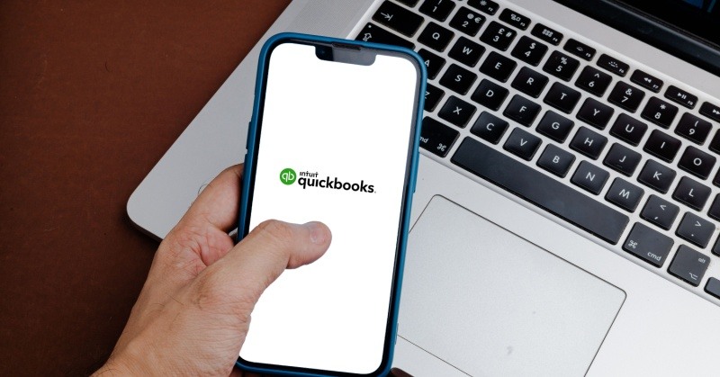 Intuit QuickBooks app on smartphone held up by hand MacBook laptop on desk in background