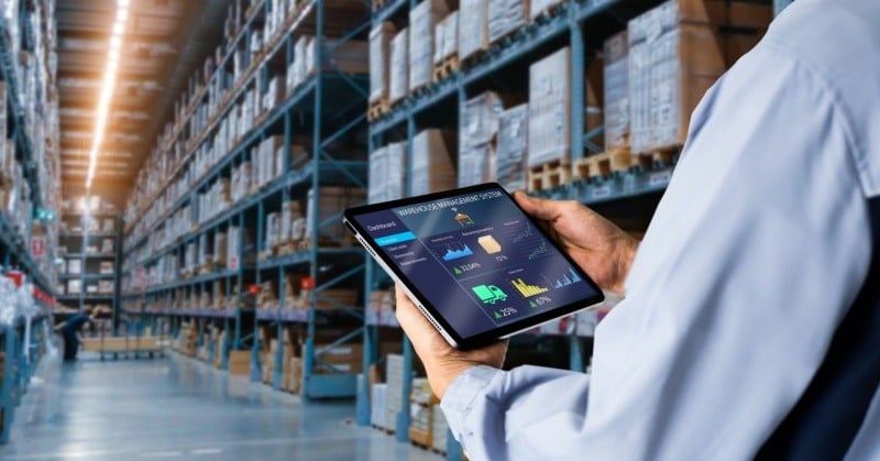 unseen person in white shirt managing inventory on tablet in warehouse full of wood pallets stacked on blue metal shelves