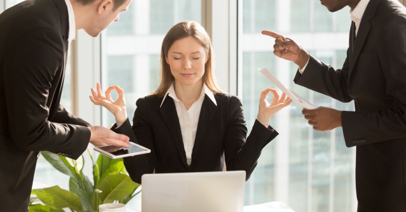 business mindfulness meditation woman in black suit seated in front of a laptop calm eyes closed while two male colleagues in black suits argue around her