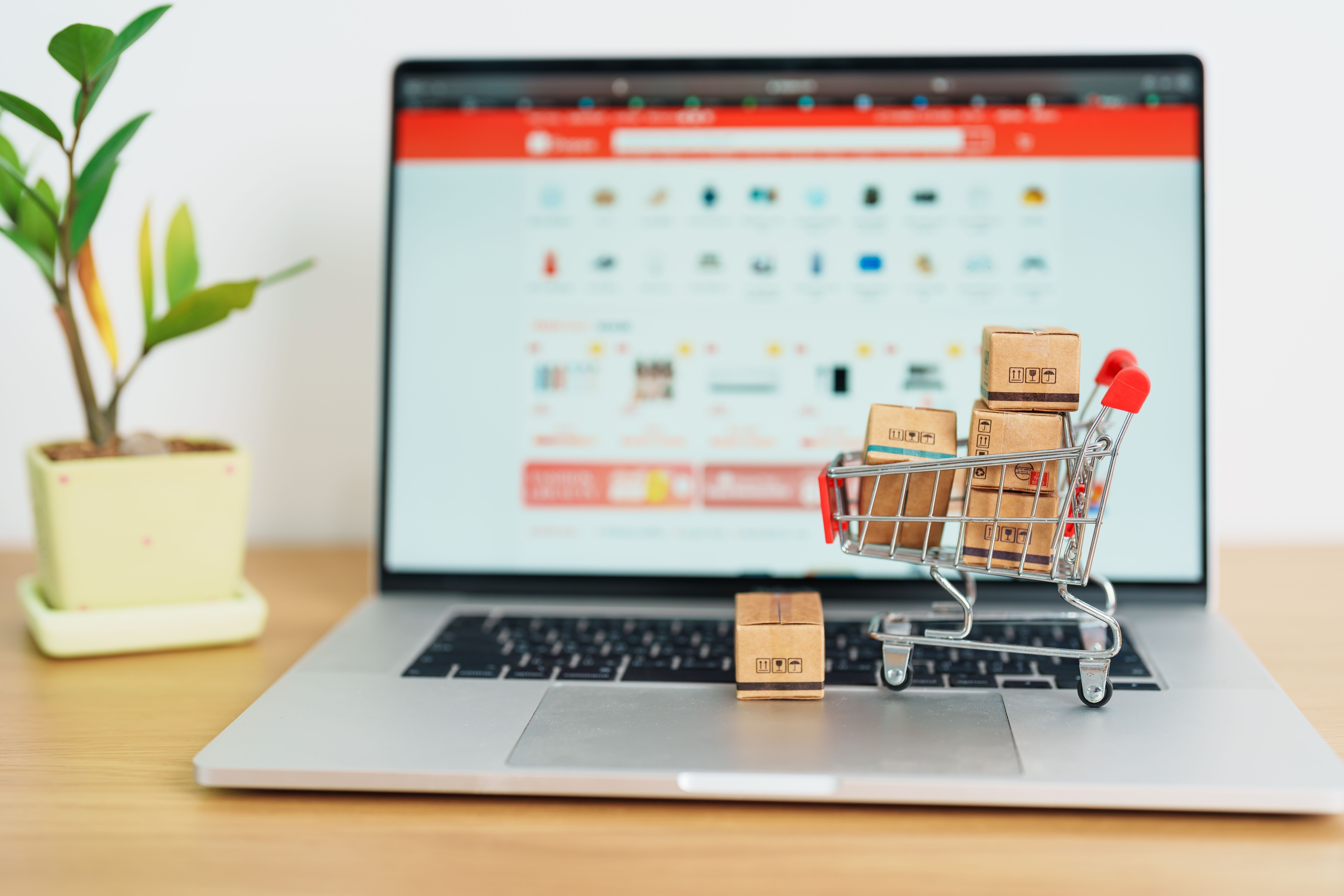 shopping cart on laptop full of tiny boxes on light brown desk with plant