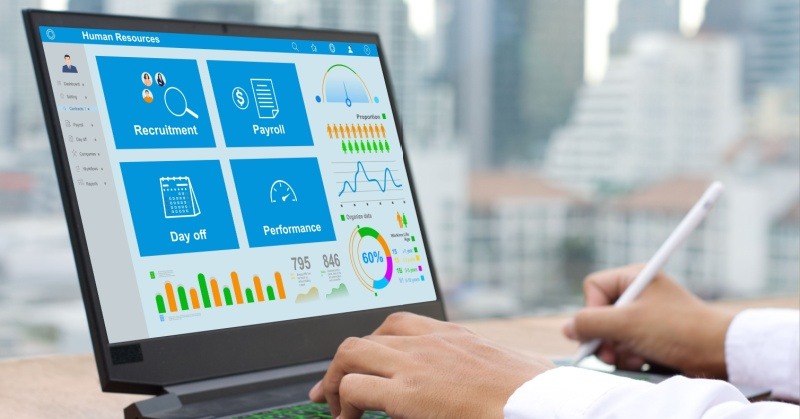 black laptop with blue icons white text that say recruitment, payroll, day off, and performance on the screen, human hands typing on keyboard white shirt sleeves stylus