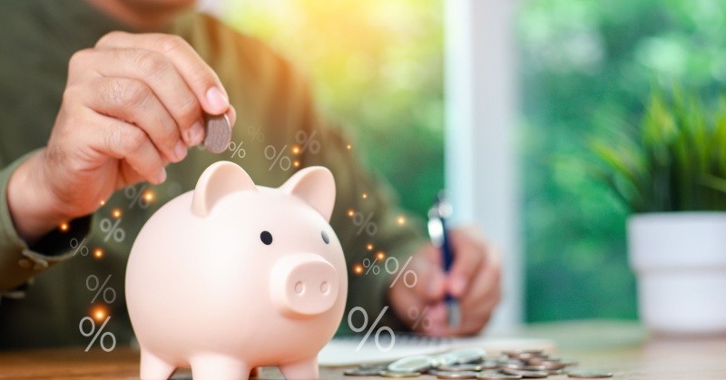 Retirement plan pink piggy bank on table next to pile of coins in front of person wearing green shirt in background writing on a piece of paper