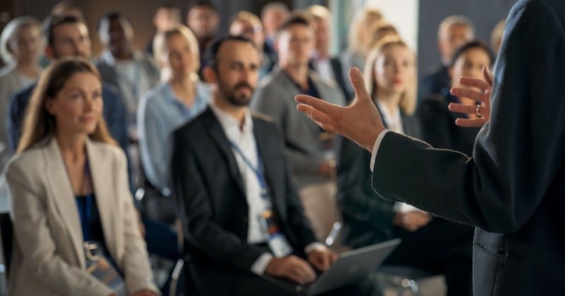 Back view of public speaker gesturing and presenting to audience of diverse business leaders male and female attendees listening to inspirational speech