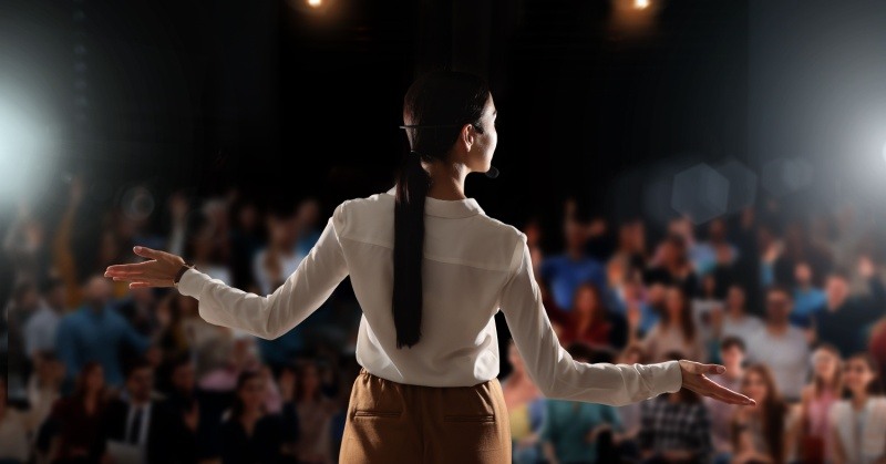 public speaker speaking to audience back to camera female long hair white shirt brown pants