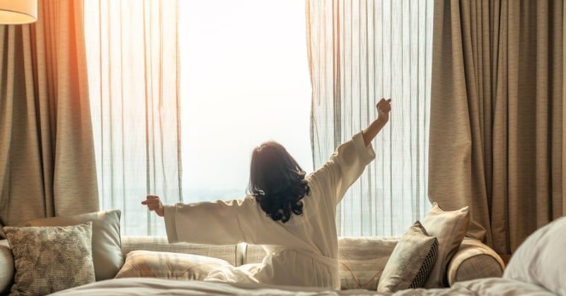 rest relaxation rejuvenation woman with dark hair in white robe stretching on bed in front of veiled window sunlight shining through